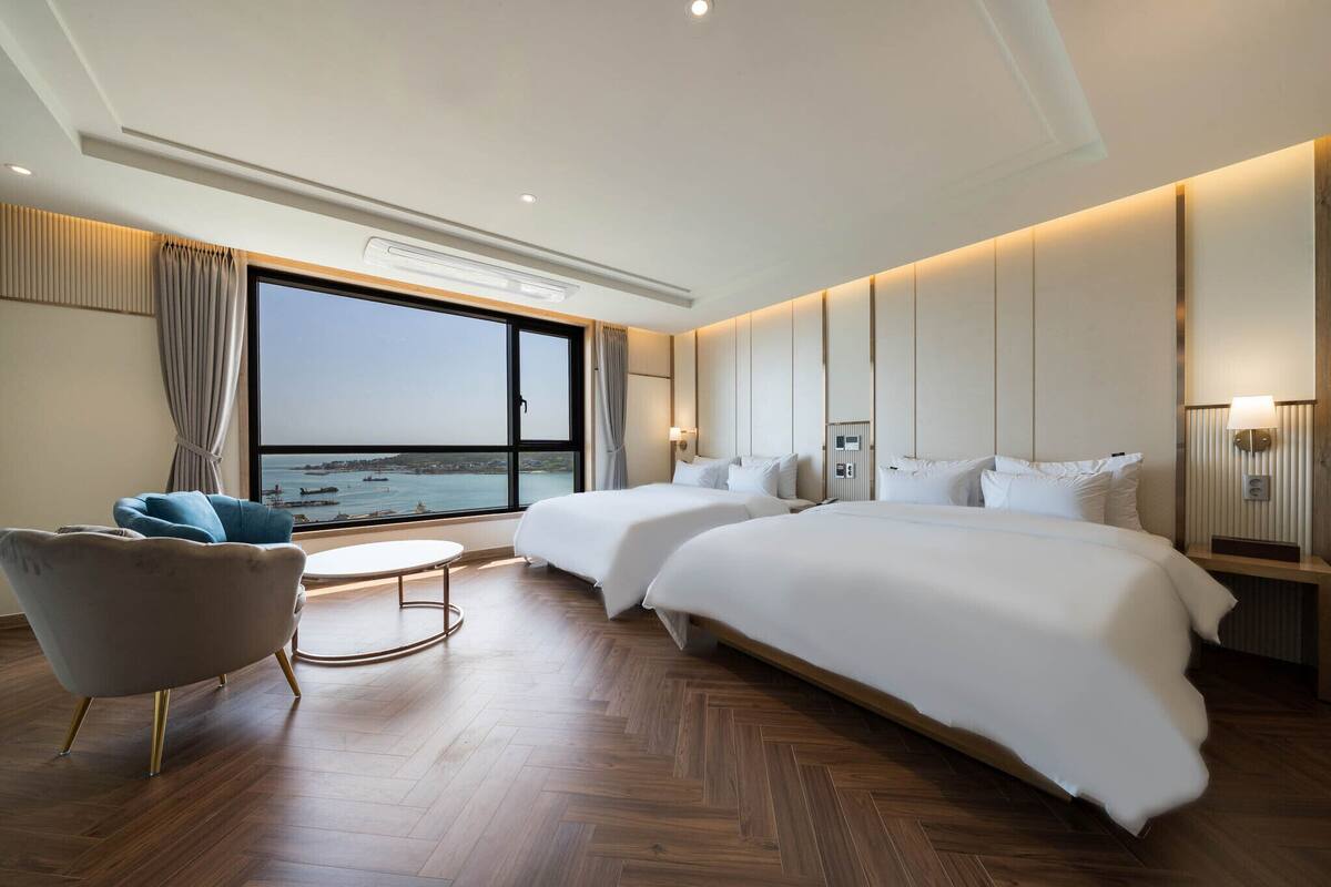 A modern hotel room with a large bed, two smaller beds, a seating area with a chair, a round table, and a view of the sea.