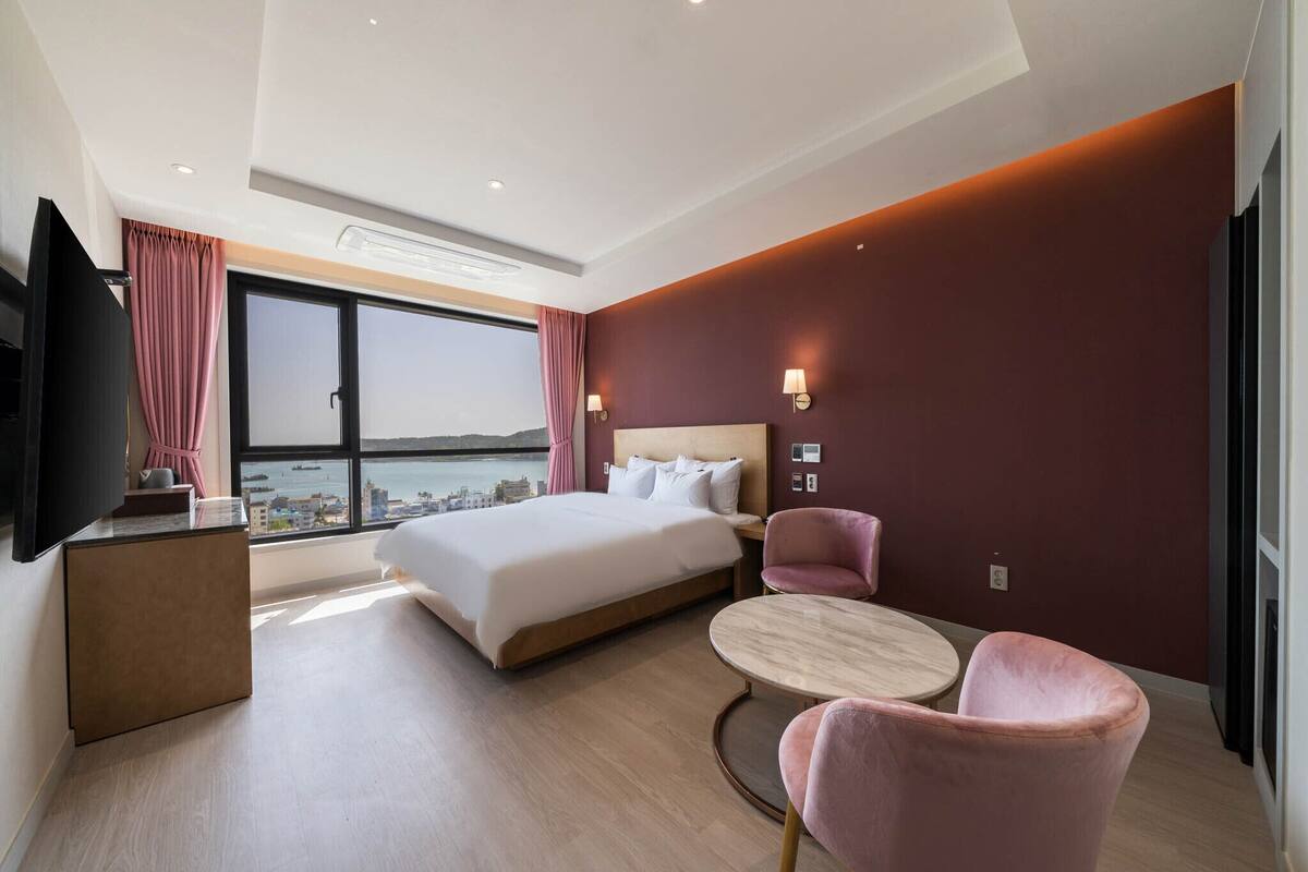 A modern hotel room with a large bed, a desk, a chair, and a view of the city through a large window.