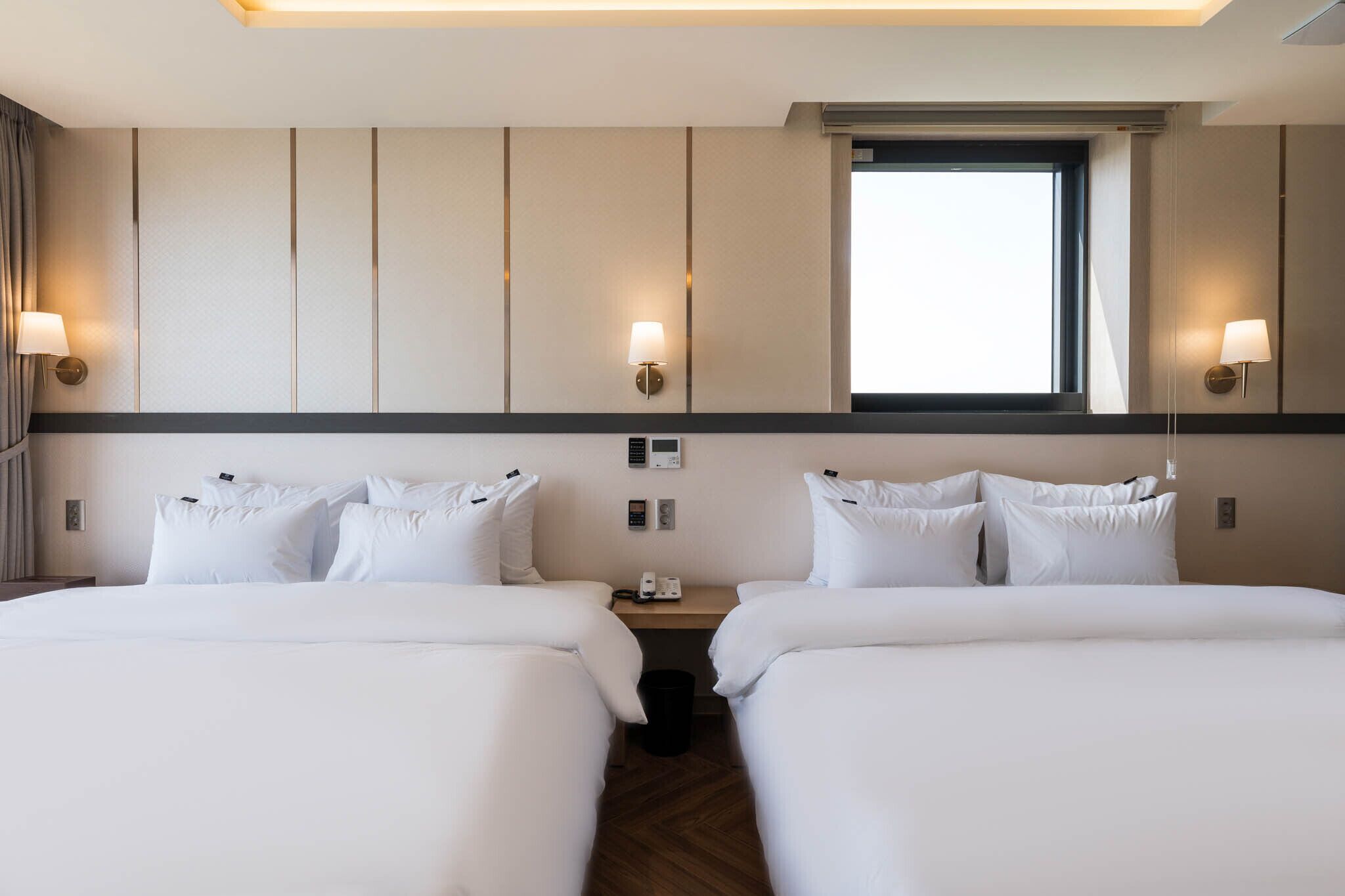 A hotel room with two beds, a window, and wall-mounted lamps.