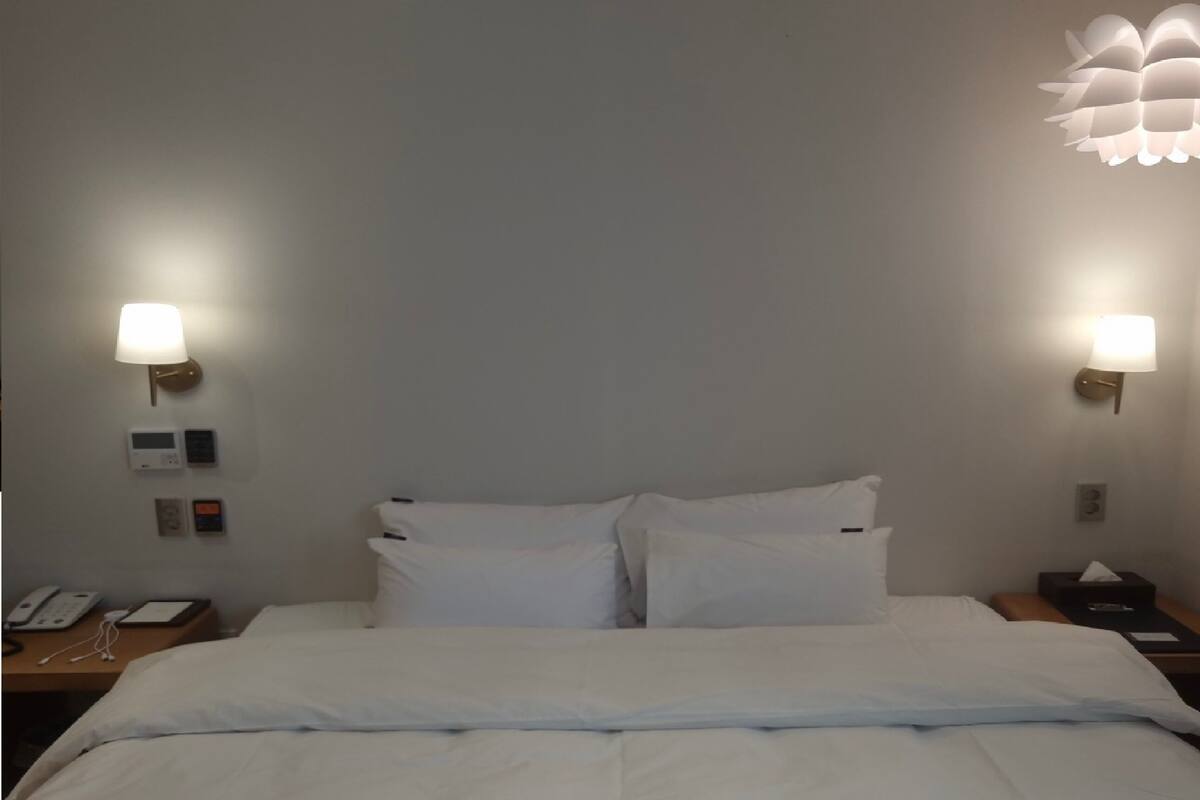 A hotel room with a bed, two bedside tables, a wall-mounted lamp, a thermostat, and a phone.
