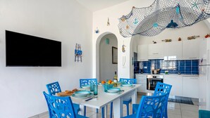 Een moderne keuken met witte kasten, een eettafel met blauwe stoelen en een aan de muur bevestigde televisie.