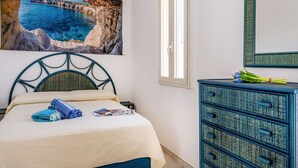 Een slaapkamer met een bed, een dressoir, een schilderij van een kustlandschap en een raam.