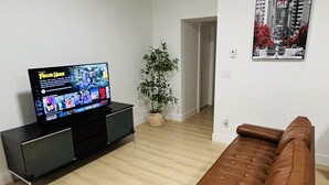 Ein Wohnzimmer mit Flachbildfernseher auf schwarzem Standfuß, braunem Ledersofa, einer Topfpflanze und einem gerahmten Stadtbild an der Wand.