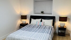 Ein Schlafzimmer mit einem Bett, zwei Nachttischen, einem Fenster mit Jalousien und einer Deckenleuchte.