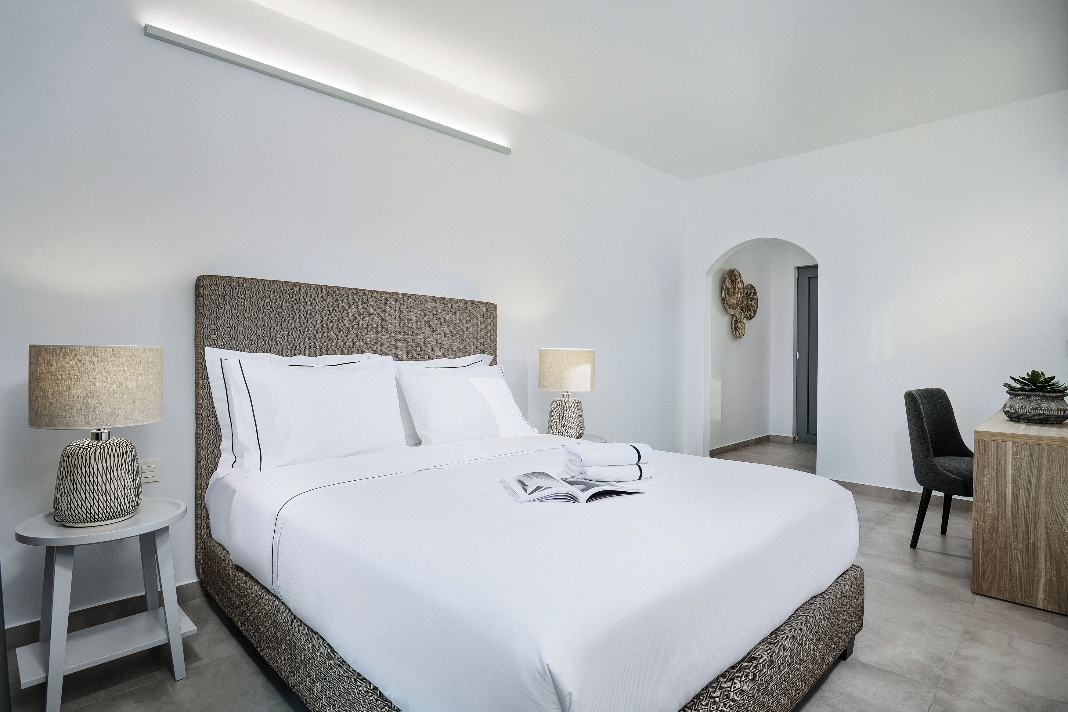 Una habitación de hotel moderna con una cama grande, dos mesitas de noche con lámparas, un escritorio pequeño y una planta en maceta.