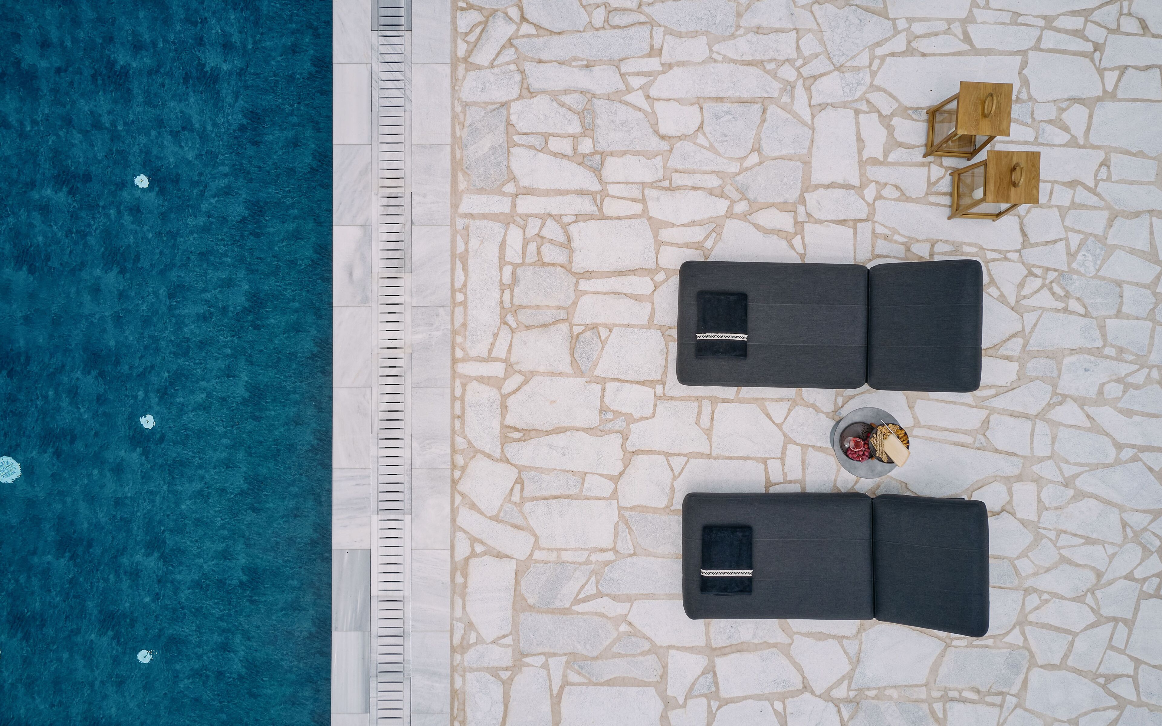 Vista aérea de uma área de piscina com duas espreguiçadeiras, uma pequena mesa com uma bebida e piso de ladrilho de pedra.