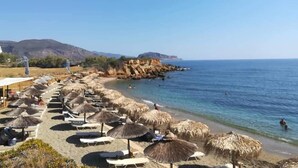 Une plage avec des chaises longues et des parasols en chaume, une mer d’un bleu limpide et, au loin, des falaises rocheuses.