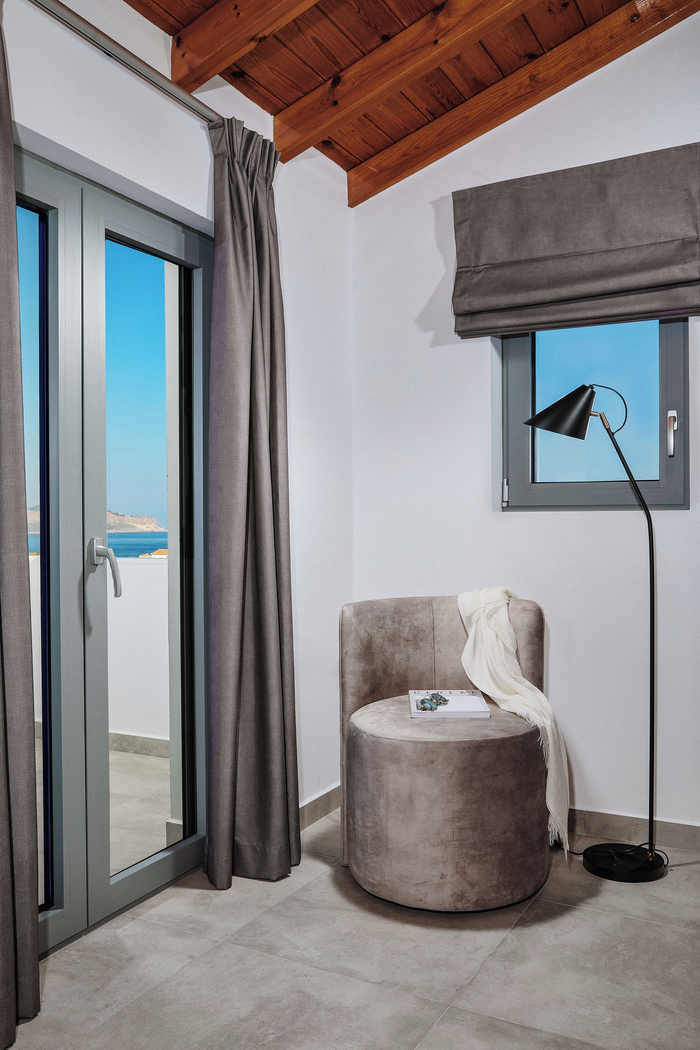 Quarto com porta de vidro deslizante para uma varanda, uma cadeira redonda com almofadas, um abajur e uma janela com vista para o mar.