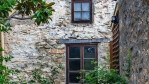 Un edificio de piedra con una ventana central y una puerta pequeña, rodeado de follaje y un camino empedrado.
