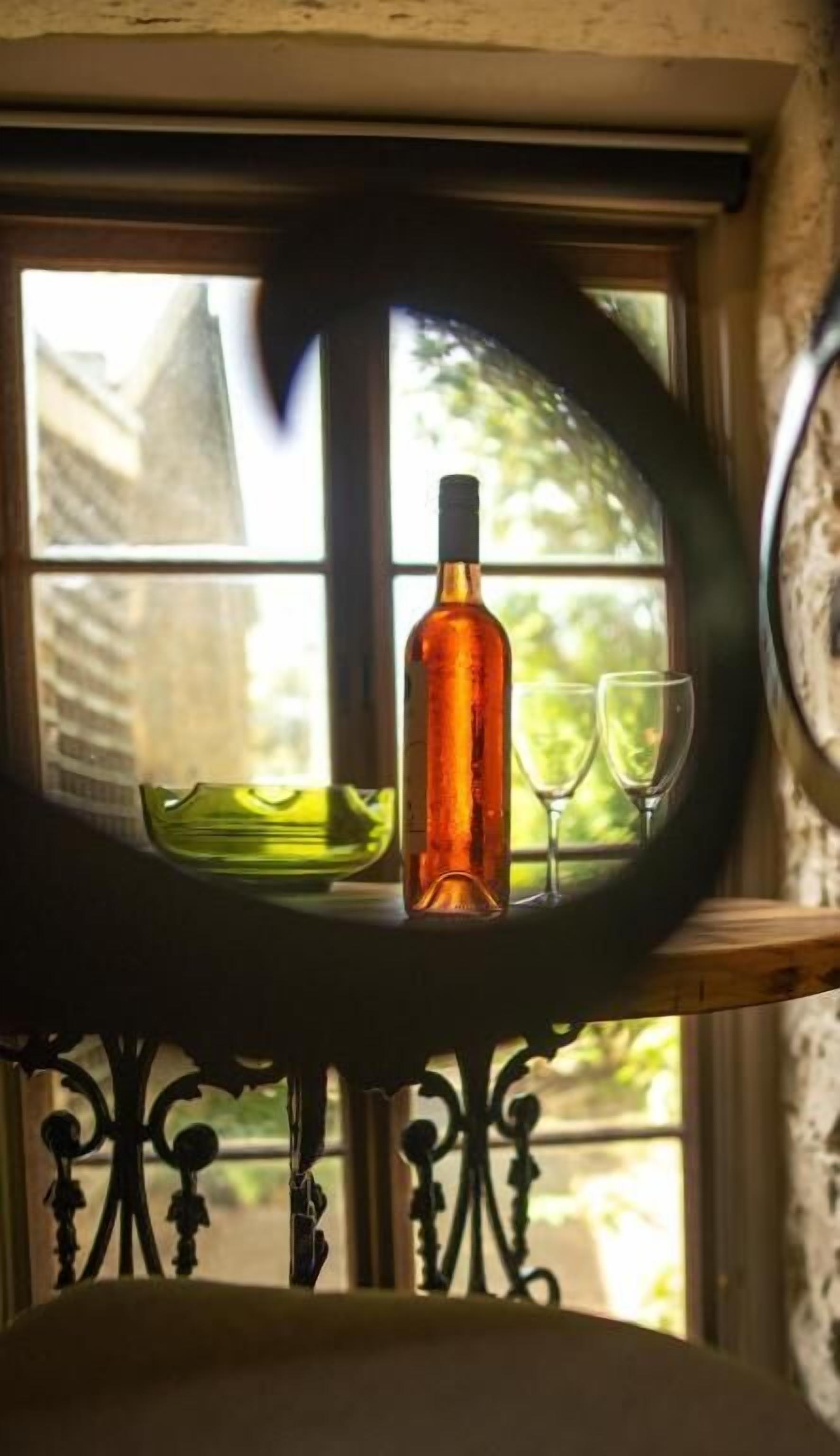 A bottle of wine and two glasses on a table by a window with a view of buildings and greenery.