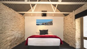 Un dormitorio con cama, manta roja, almohada negra, pared de piedra y un cuadro enmarcado de una playa.