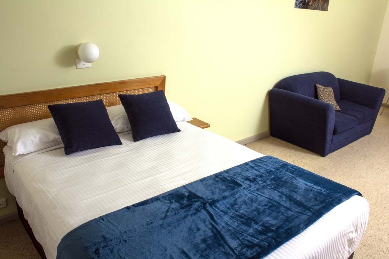 Una cama con una colcha blanca y azul, dos almohadas azul oscuro y un cabecero de madera. Hay un sillón azul oscuro con un cojín y una lámpara blanca montada en la pared.
