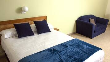 Una cama con una colcha blanca y azul, dos almohadas azul oscuro y un cabecero de madera. Hay un sillón azul oscuro con un cojín y una lámpara blanca montada en la pared.