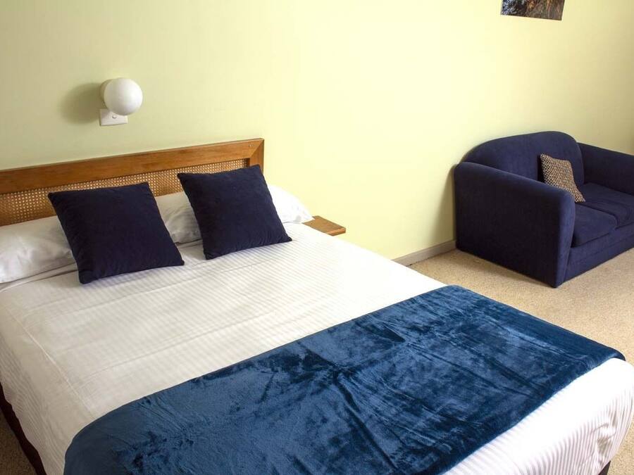 Una cama con una colcha blanca y azul, dos almohadas azul oscuro y un cabecero de madera. Hay un sillón azul oscuro con un cojín y una lámpara blanca montada en la pared.