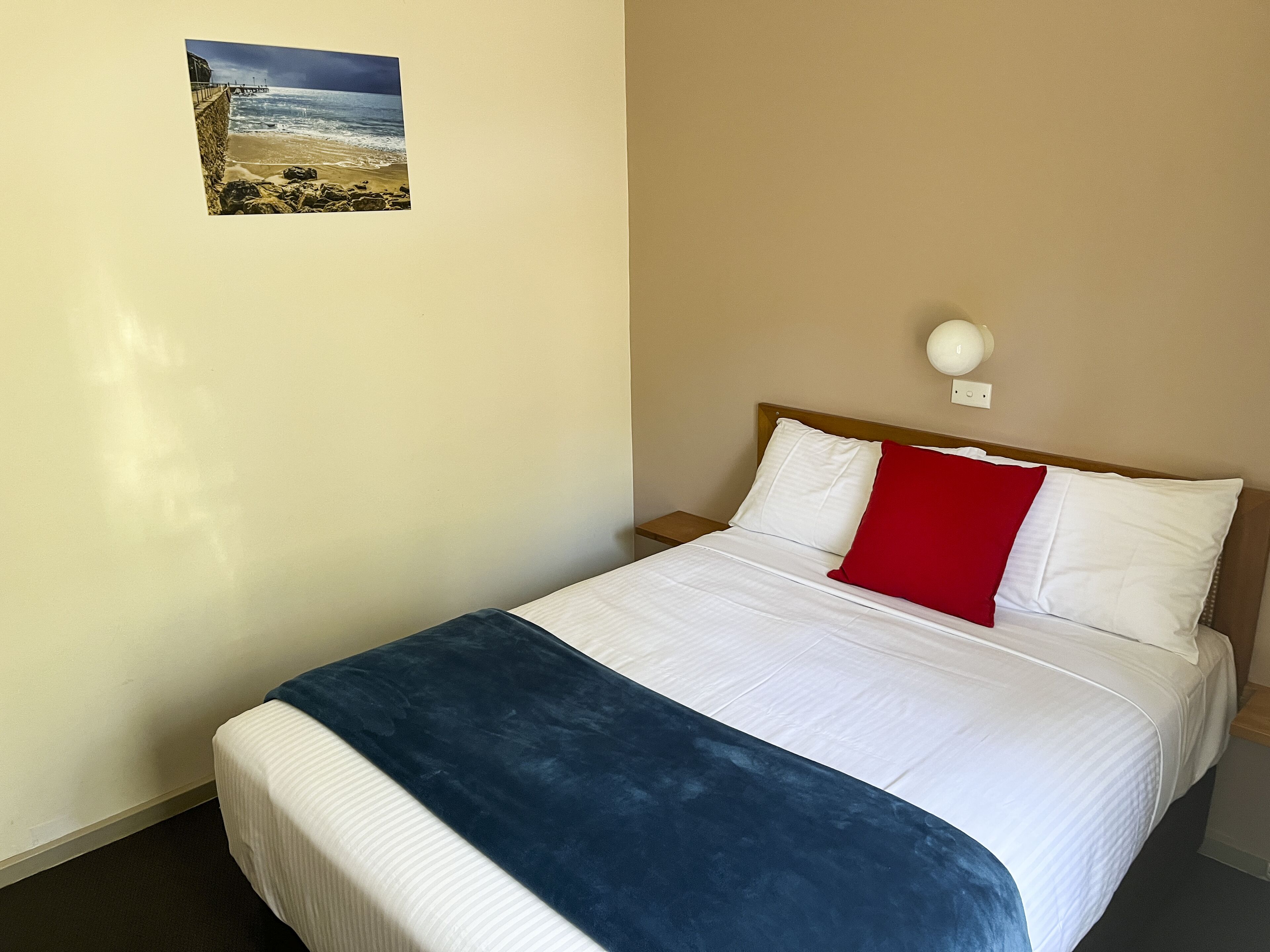 A hotel room with a bed, a nightstand, a wall-mounted lamp, and a picture of a beach scene.
