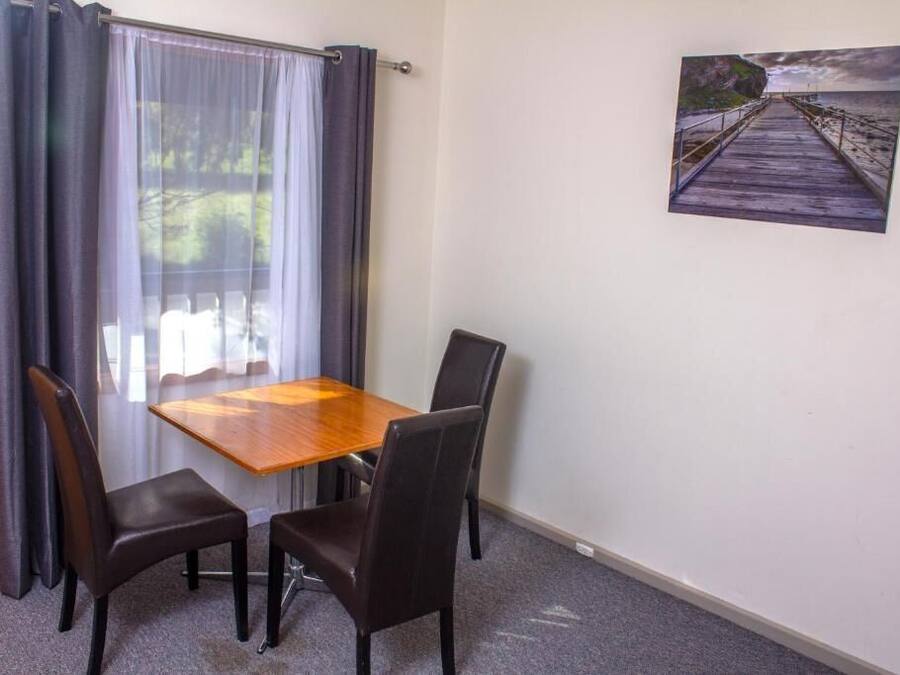 A room with a wooden table and four chairs, a window with curtains, and a painting of a pier on the wall.
