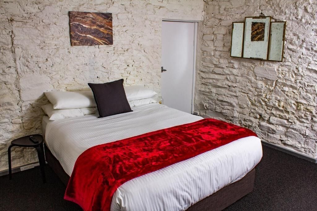 A bedroom with a stone wall, a bed with white bedding and a red throw, a black pillow, and a small stool.