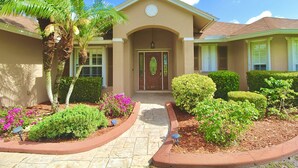 A two-story house with a front entrance, a driveway, and well-maintained landscaping including a palm tree, shrubs, and flowering plants.