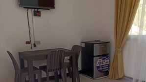 Une pièce comprenant une table à manger, des chaises, un téléviseur fixé au mur, un réfrigérateur et une fenêtre avec des rideaux.