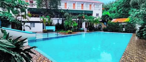 Una piscina con borde de azulejos, rodeada de vegetación y un edificio con techo rojo.