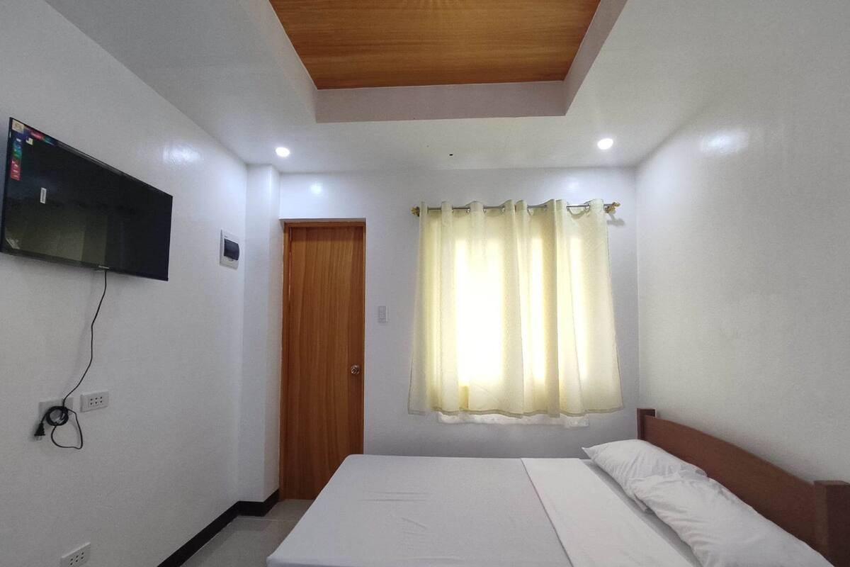A bedroom with a bed, a TV mounted on the wall, a wooden door, and a window with curtains.
