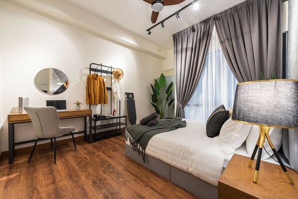 Un dormitorio moderno con suelo de madera, cama con edredón gris y blanco, escritorio con computadora, una silla, un espejo y una planta grande.