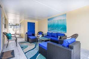 A sunlit patio with wicker furniture, a blue door, and a large wave painting on the wall.