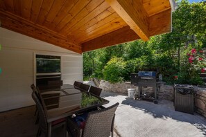 Eine Terrasse im Freien mit Holzboden, einem Esstisch mit Stühlen, einem Grill und einer Steinwand mit Bepflanzung.