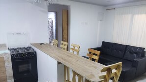 A compact living space with a kitchenette, a dining table, wooden chairs, a black sofa, and a trash bin. - San San Andrés Magic House 0 by BBBSAI (San Andrés y Providencia)