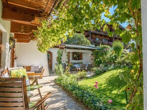 Un edificio in legno rustico con portico coperto, pavimento in pietra e un giardino con diverse piante e fiori.