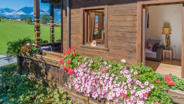 Un chalet de madera con balcón, una ventana con espejo y vistas a un campo verde y montañas.