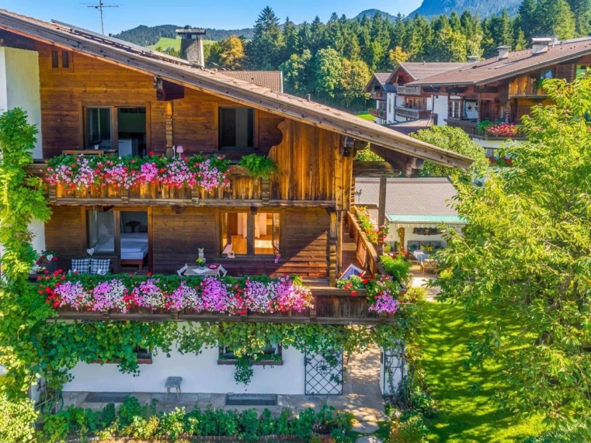 A two-story wooden chalet with balconies adorned with flowers, set in a mountainous area.