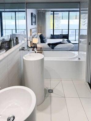 Une salle de bain moderne avec un grand lavabo, une baignoire et une douche. La pièce est dotée d’une grande fenêtre offrant une vue sur la ville.