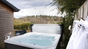 Een jacuzzi in een achtertuin met een houten huis, een fles champagne en handdoeken aan een haak.