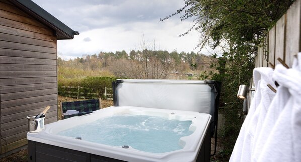 A hot tub in a backyard with a wooden house, a bottle of champagne, and towels hanging on a hook.