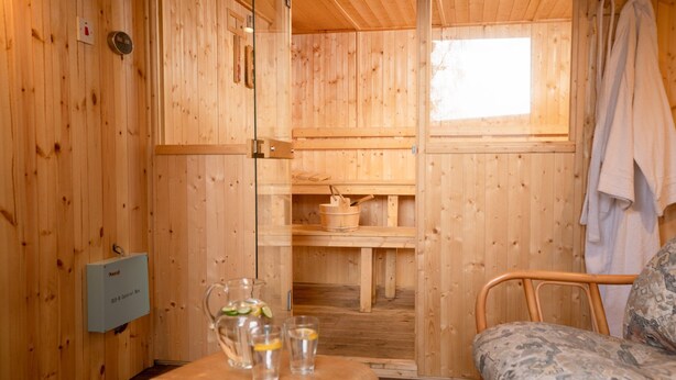 Een houten hutje van binnen, met een houten bed, een tafel met glazen en een karaf, en een raam met een handdoek erover.