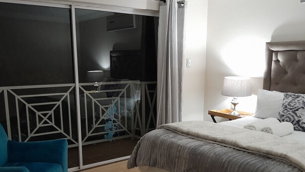 Ein Schlafzimmer mit Bett, Nachttisch, Lampe, Stuhl und Blick auf einen Balkon.