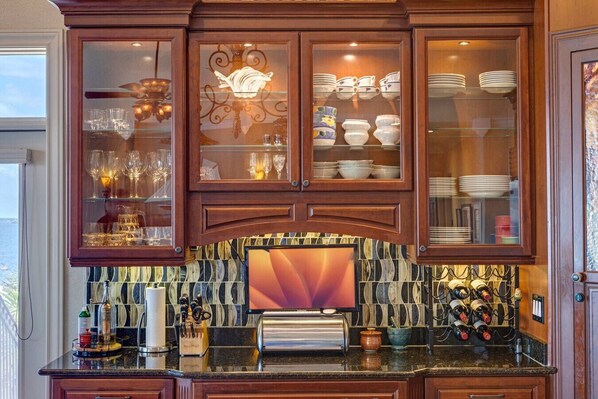 A kitchen with wooden cabinets, glass-fronted shelves displaying dishes and glasses, a tiled backsplash, and a wine rack.