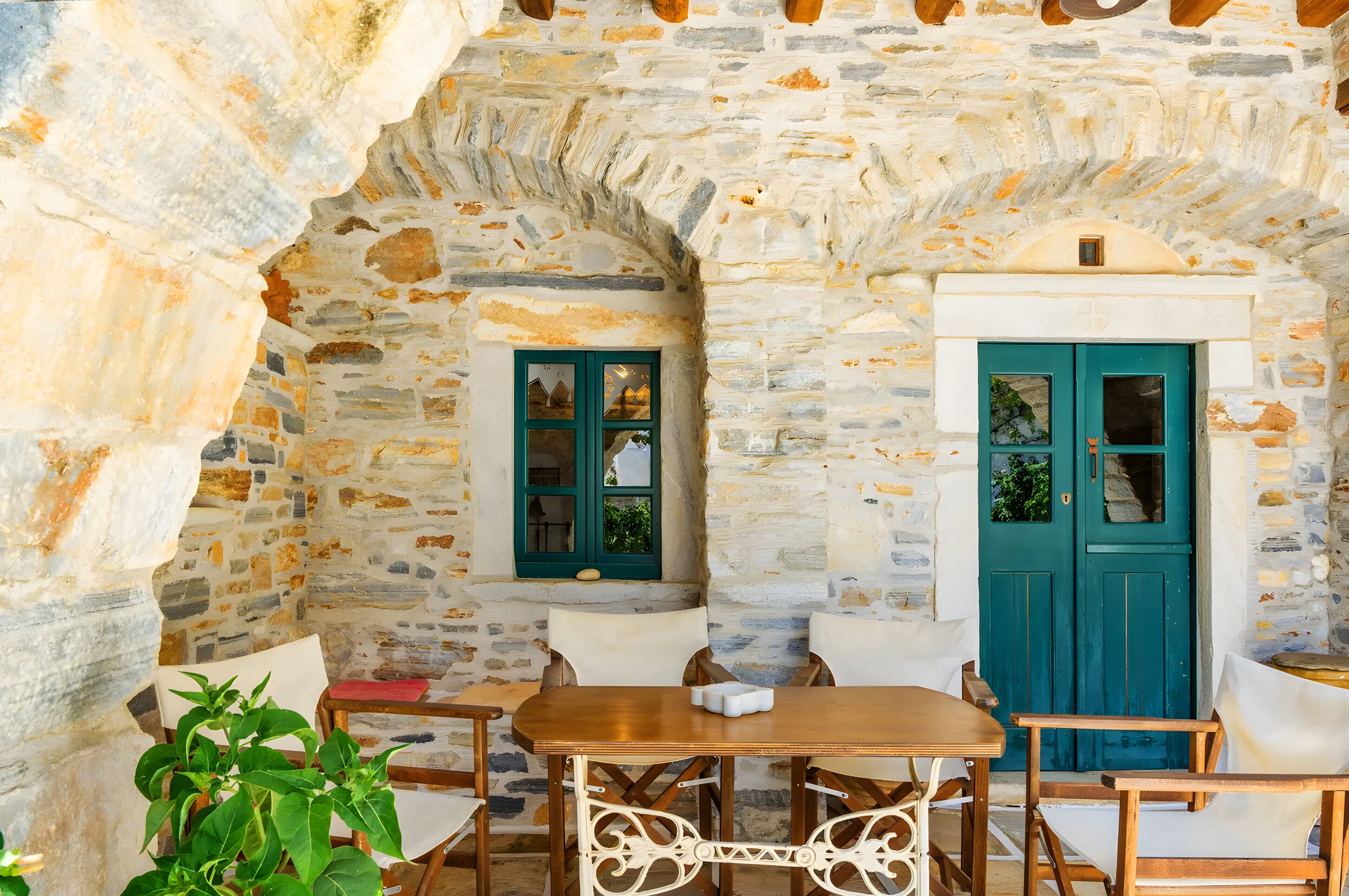 Une pièce aux murs de pierre, avec une table et des chaises en bois, une porte de couleur turquoise et une fenêtre dotée de volets verts.