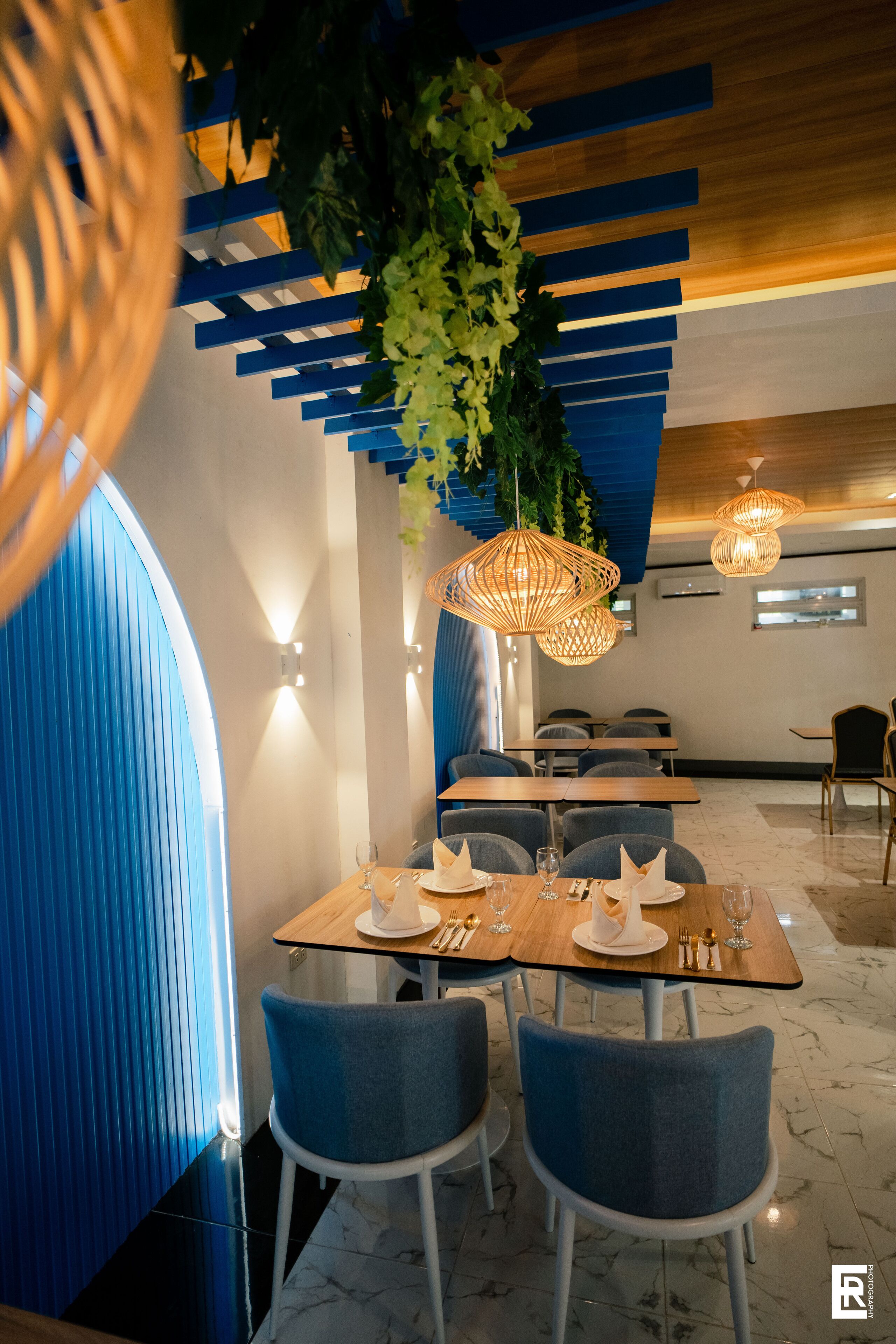 A modern restaurant interior with blue and wooden accents, marble flooring, and hanging wicker lamps.