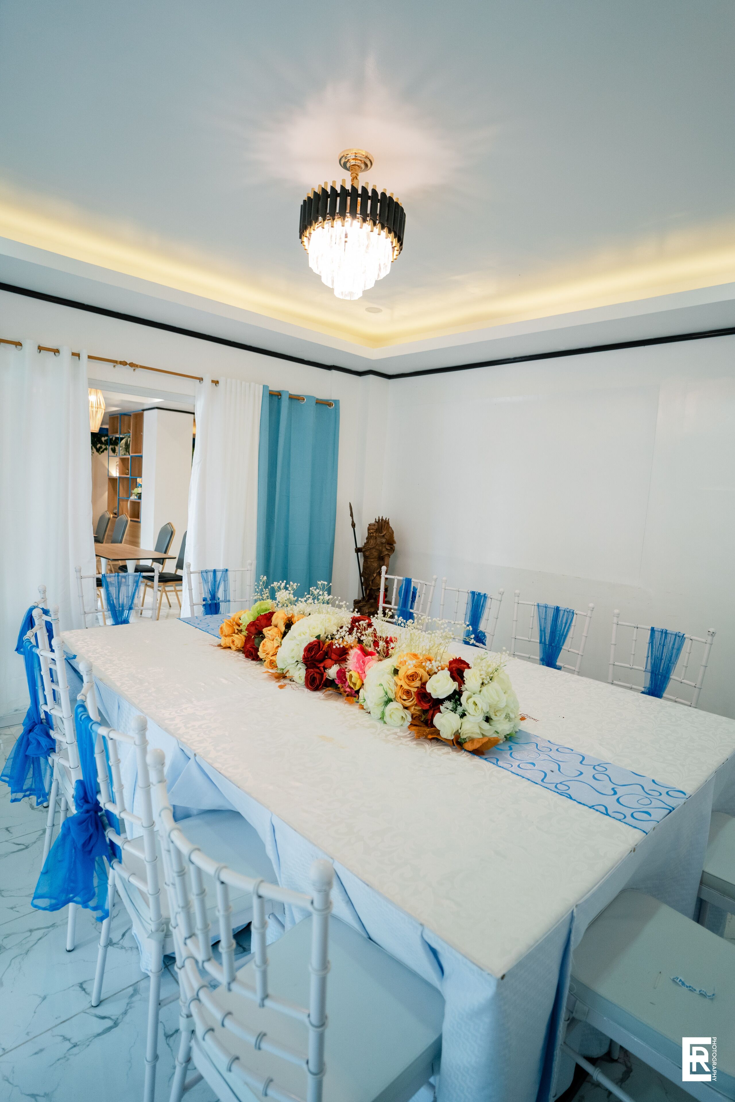 A long dining table with a floral centerpiece, white chairs with blue sashes, and a chandelier.