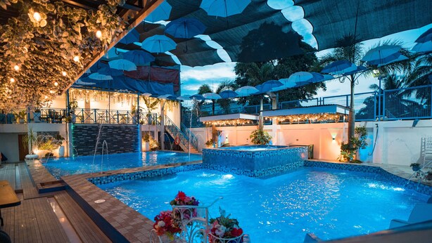 A swimming pool with a wooden deck, surrounded by umbrellas and plants, and a staircase leading to an upper level.
