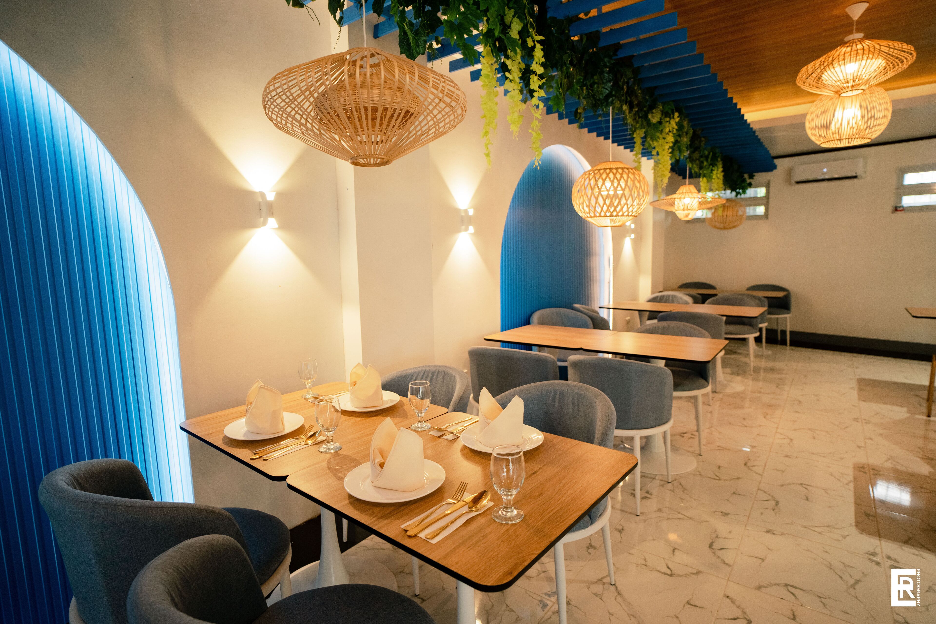 A modern restaurant interior with wooden tables, grey chairs, and hanging wicker lamps.