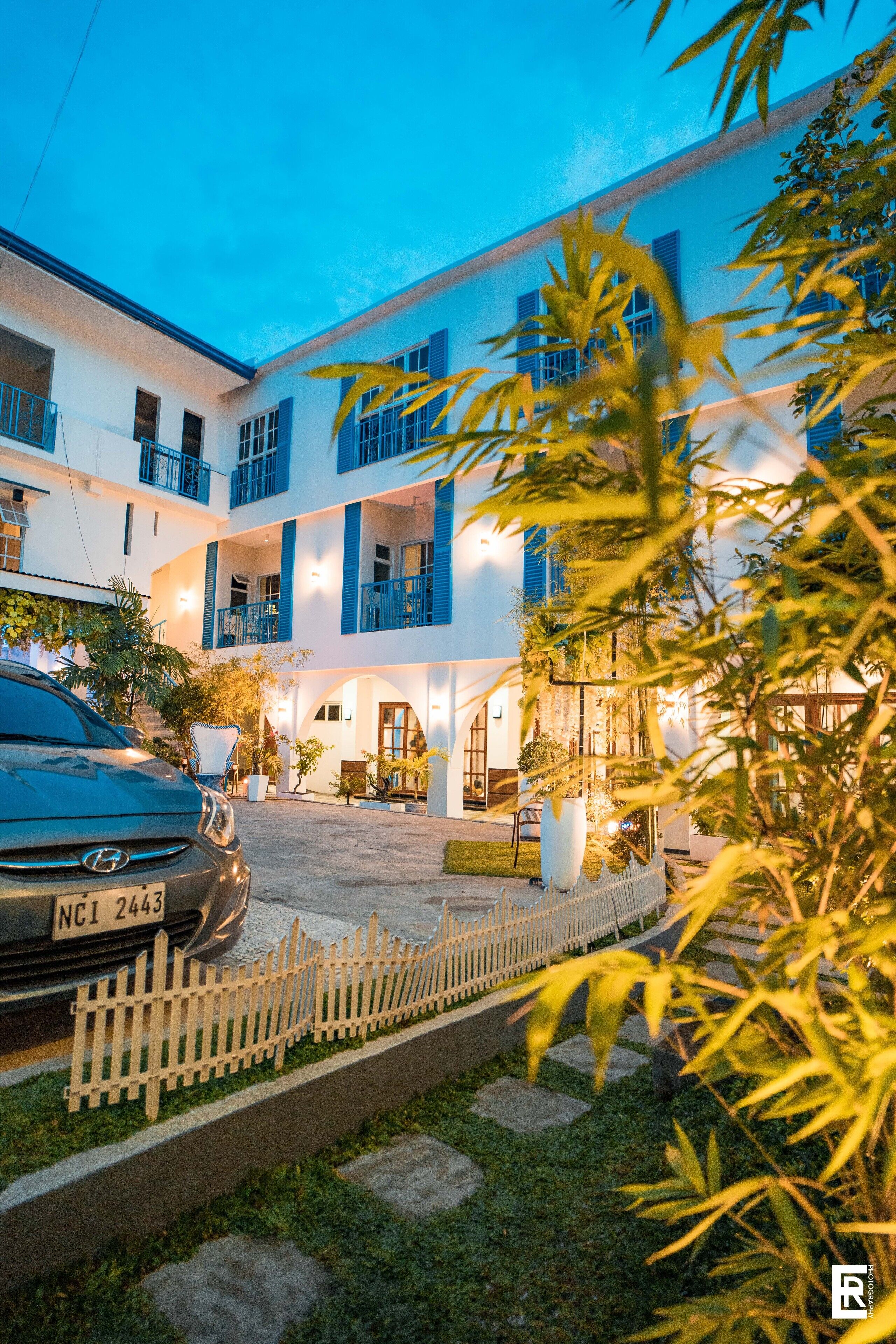 Un hotel de varios pisos con fachada blanca y detalles azules, que cuenta con un área de estacionamiento, un coche y una cerca blanca de piquetes.