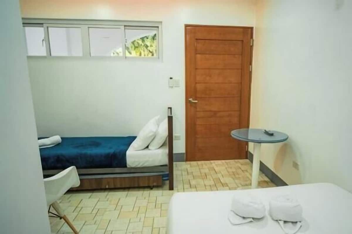 Una habitación pequeña y limpia, con una cama, una silla, una mesita y una puerta.