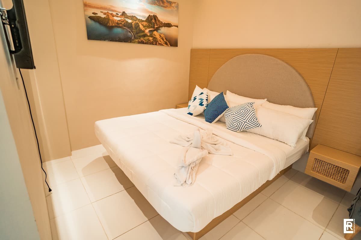 Una cama bien hecha con sábanas blancas y almohadas estampadas, un cabecero de madera y una mesita de noche con lámpara.