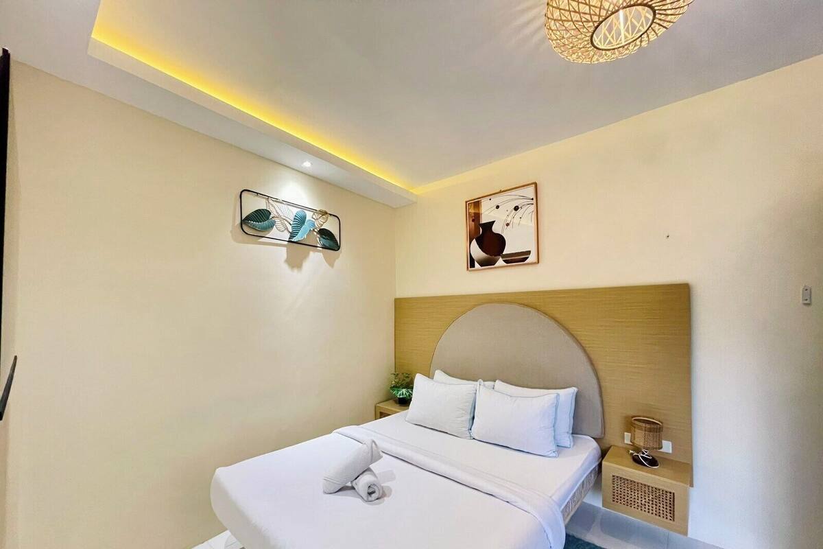 Una cama bien tendida con sábanas blancas y una toalla enrollada, un cabecero de madera, una mesita de noche con una lámpara y dos obras de arte colgadas en la pared.
