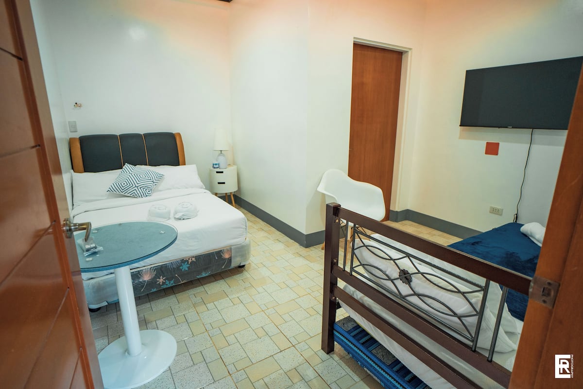 Habitación de hotel pequeña con una cama individual, una mesita redonda, una lámpara de noche y un televisor fijado a la pared.