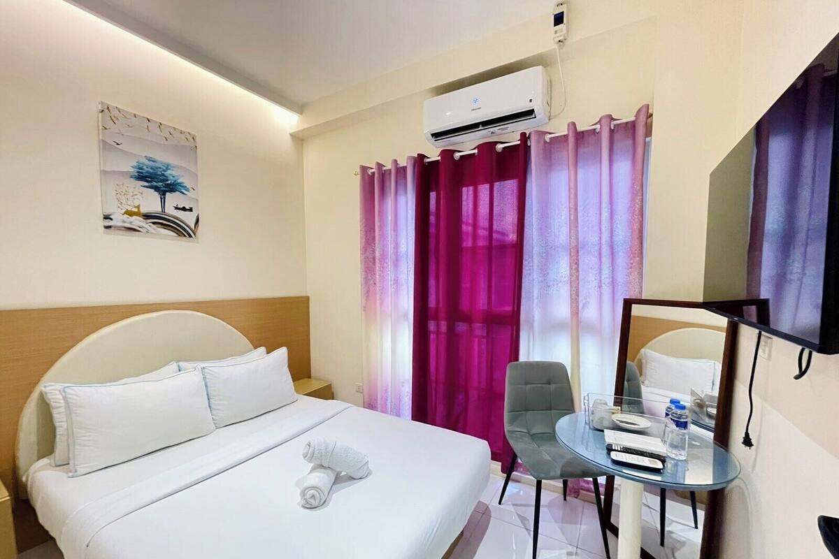 Habitación de hotel con cama, escritorio con silla, unidad de aire acondicionado, televisor y ventana con cortinas.