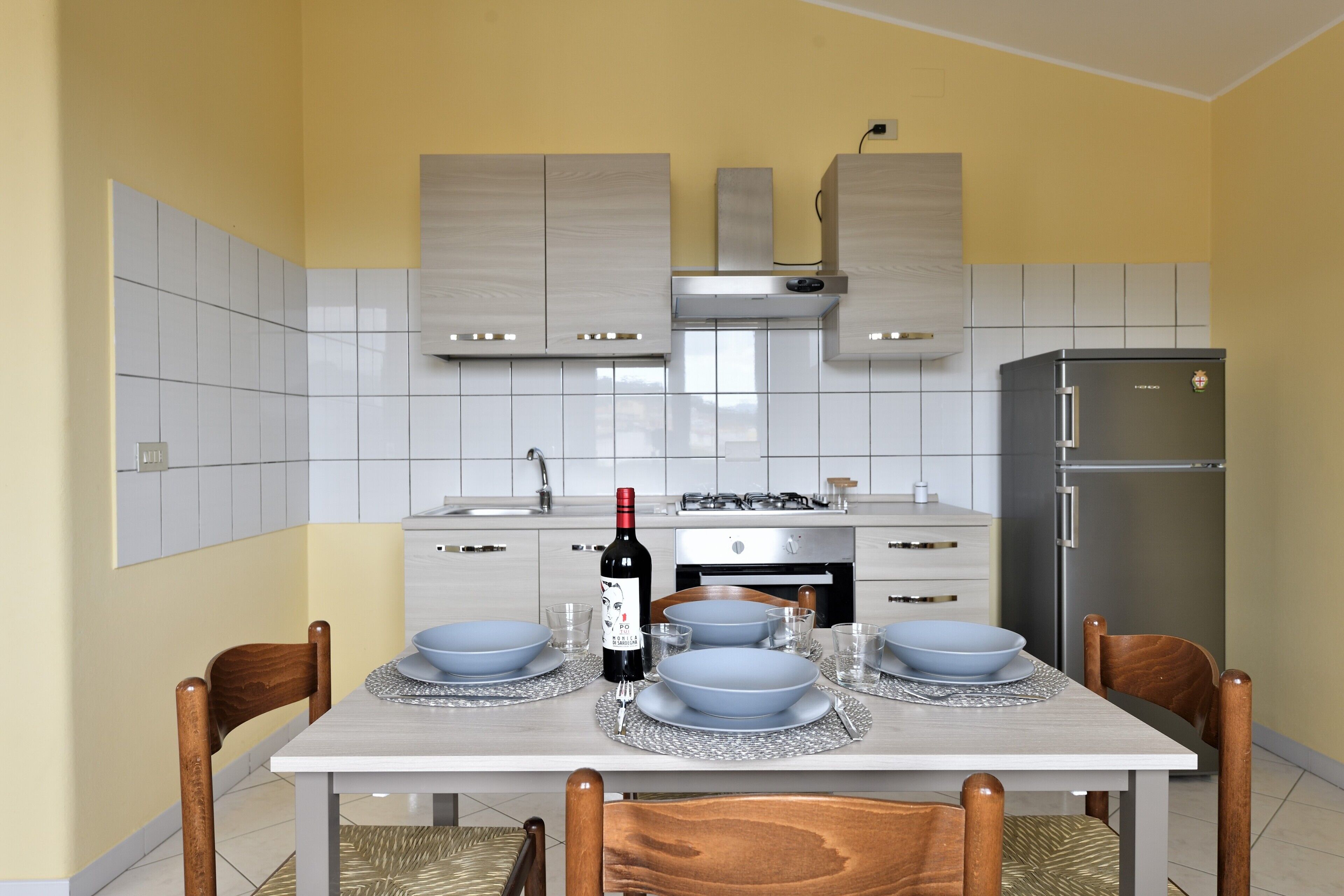 A modern kitchen with white cabinets, a stainless steel refrigerator, and a dining table set with blue plates and a wine bottle.
